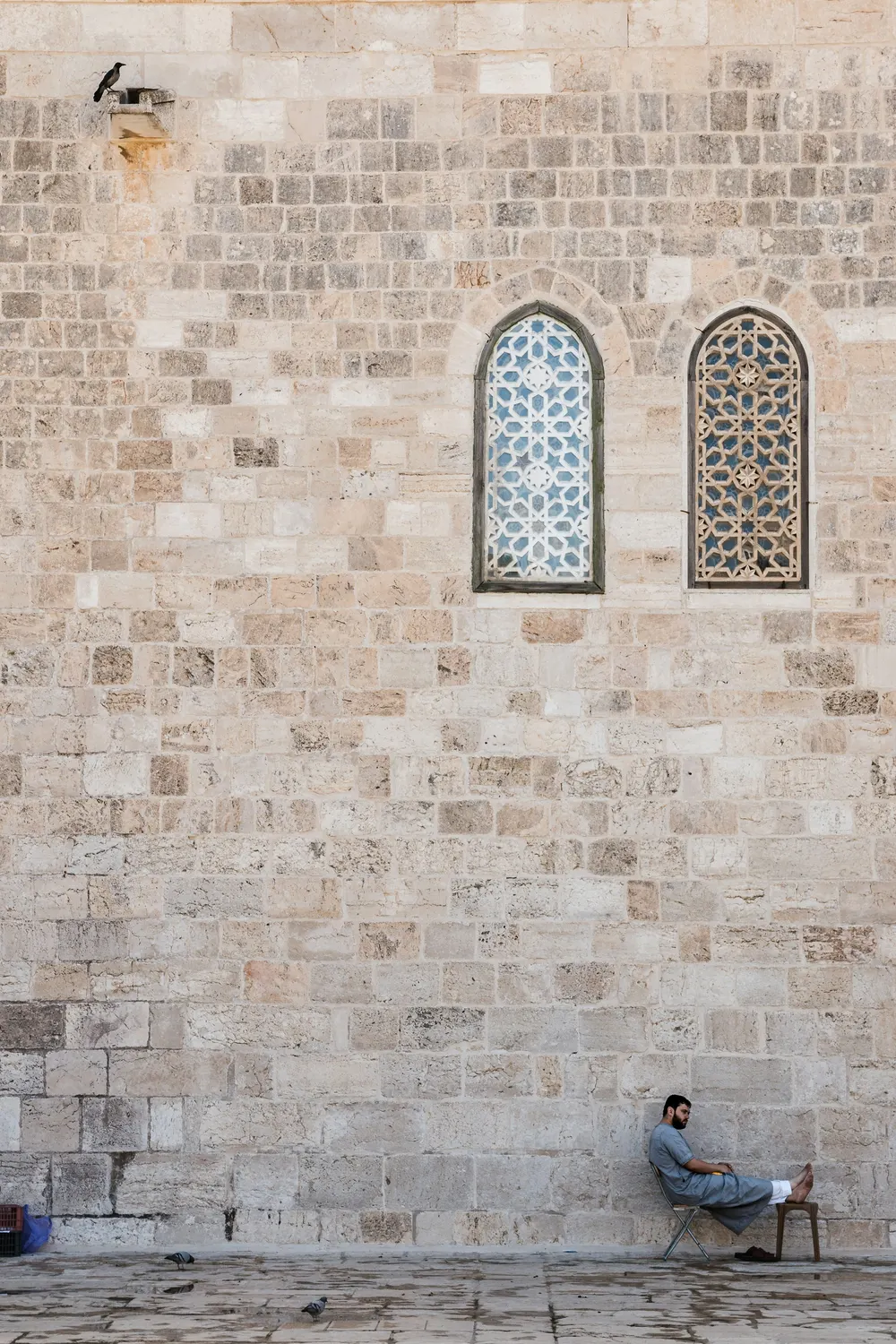 קיר אבן ירושלמית עם שני חלונות, ואיש יושב על כיסא למטה